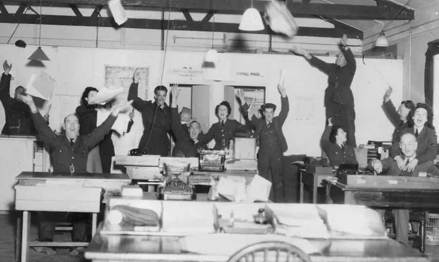 Orderly room staff at RAF Andover celebrate victory in Europe, May 1945. A black and white photograph showing RAF staff celebrating in an office, throwing their arms and papers in the air.