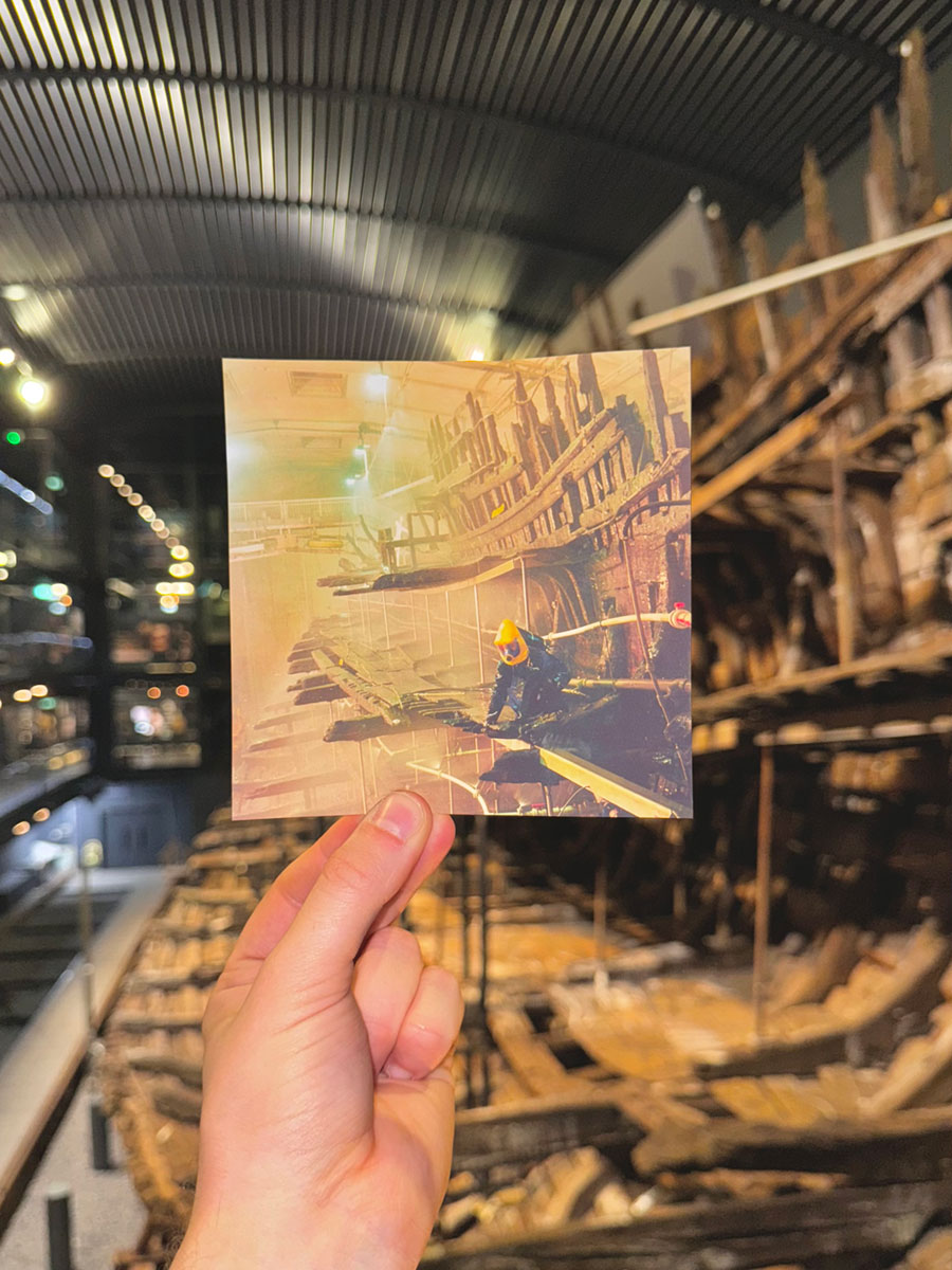 A hand holding a photograph of a person wearing a protective suit among the wreck of a ship, in front of that same wrecked ship