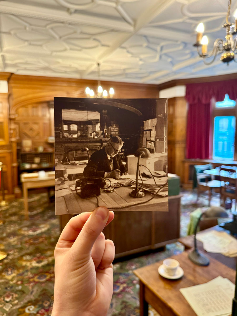 A hand holding a photograph of a man sitting at a desk, in front of that same desk in an office 