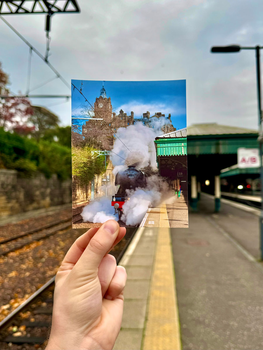 A hand holding a photograph of a train gushing steam as it prepares to pull away from the station, on the platform of that same station