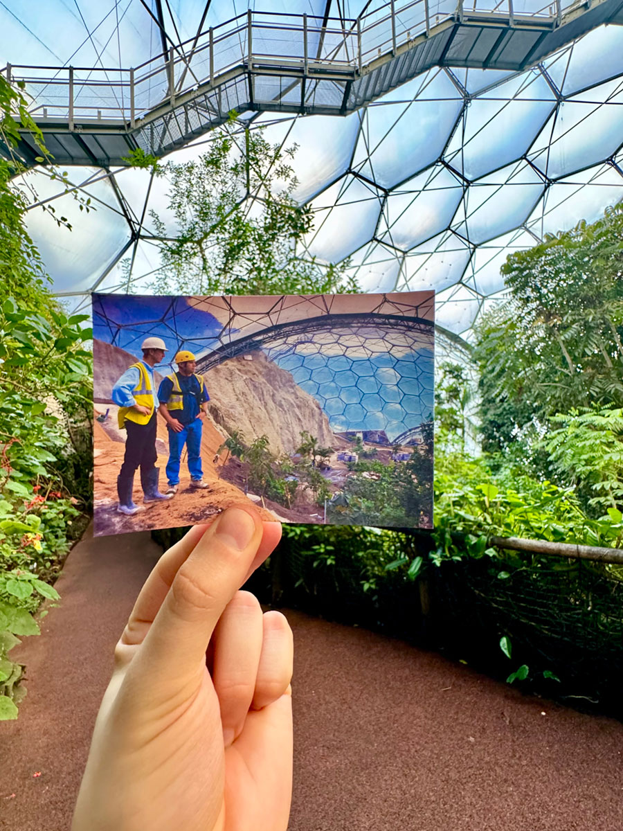 A hand holding a photograph of two people standing inside a geodesic dome, with that same geodesic dome in the background