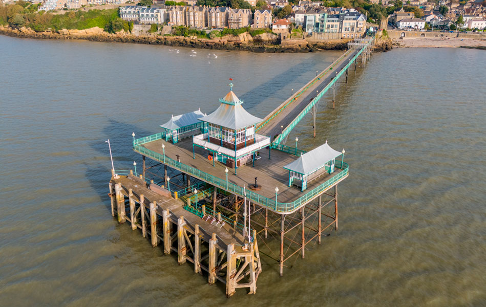 A pier with three buildings on its end, viewed from above, looking back towards the shore.