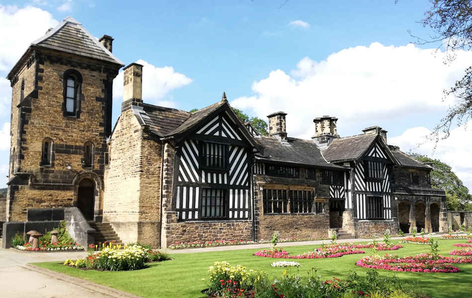 A large brick building with some Tudor-style features. In the foreground are gravel paths and a lawn with flowers planted in it.