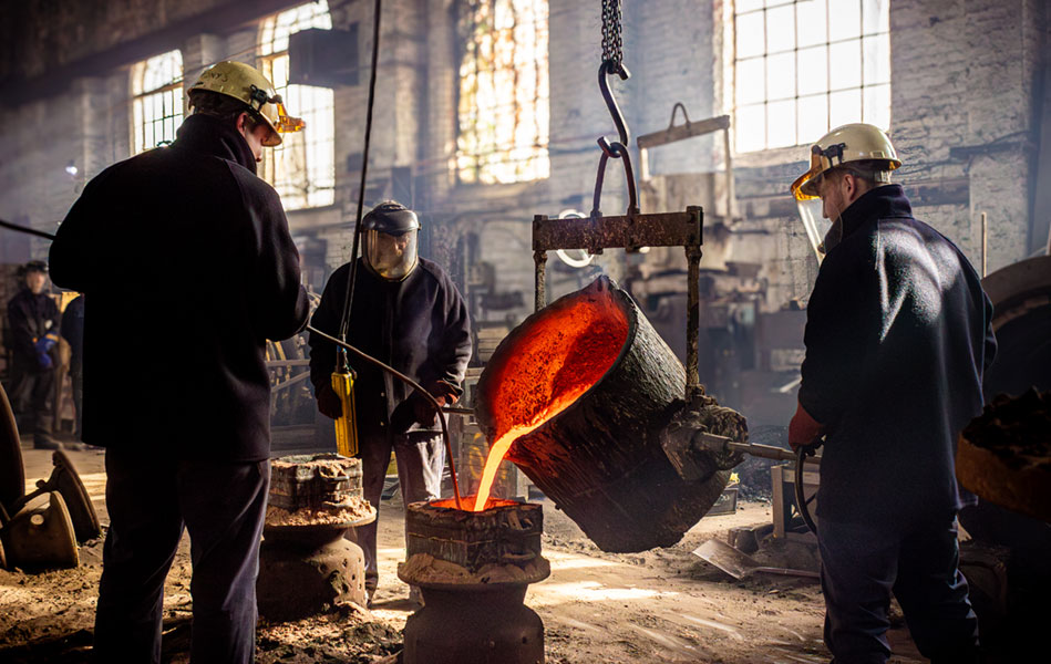 Three men in a foundry wearing safety equipment while pouring red hot metal into a bell mould.