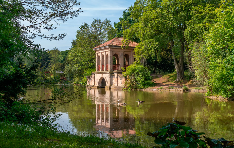 A lake with a small classical-style structure on its edge, surrounded by trees and other green vegetation.