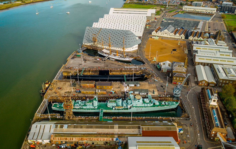 An aerial view over an industrial site featuring several large boats and buildings, situated alongside a river.