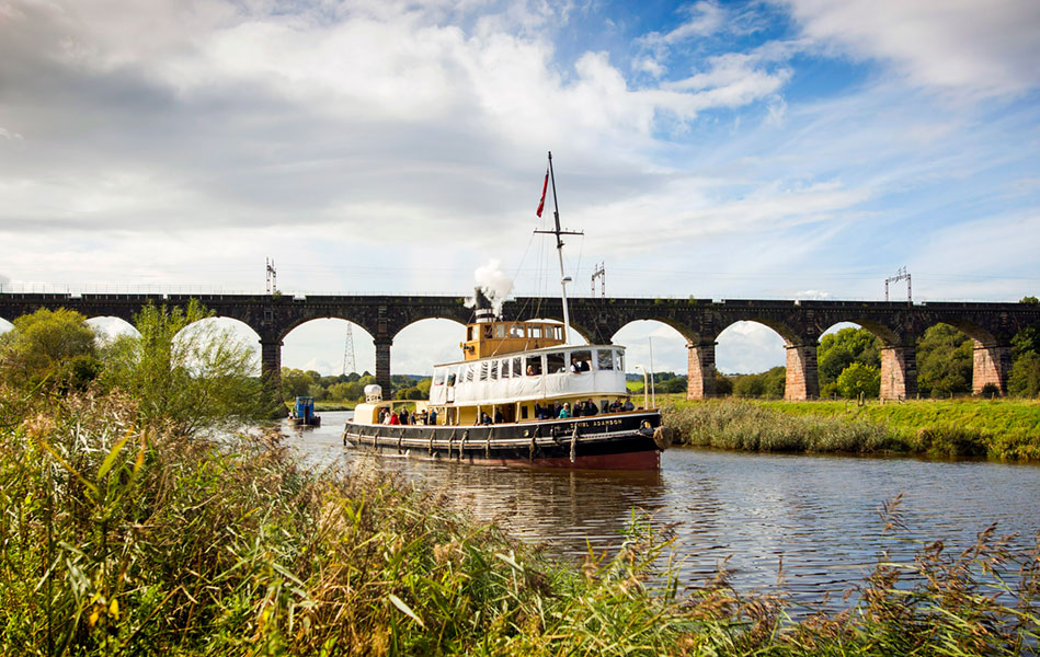 A steam powered tug boat sailing down a canal. There is a bridge in the background and green vegetation on both sides of the riverbank.