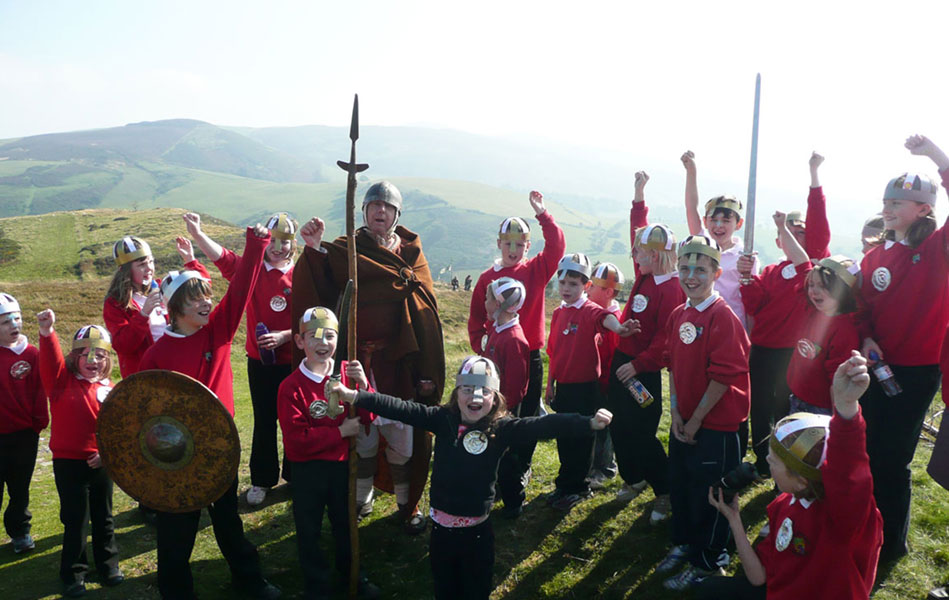 A group of schoolchildren and a man dressed in Iron Age clothing, standing in a hilly landscape. They are holding replica spears and shields and raising their hands in celebration.