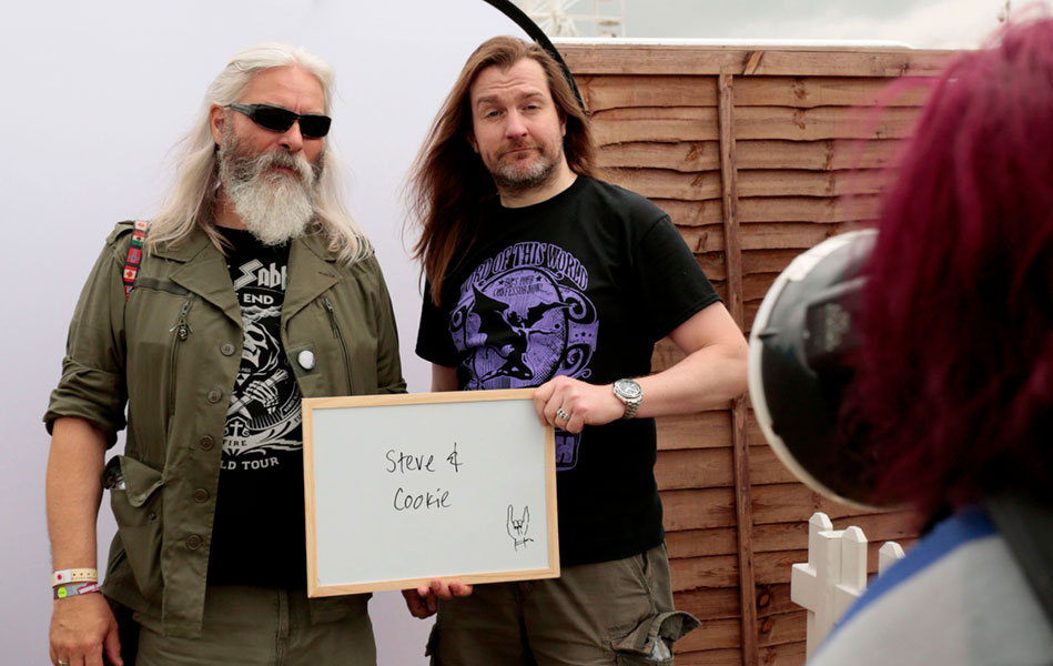 Two men wearing heavy metal band T-shirts holding a small whiteboard with their names on it, being photographed in front of a white backdrop.