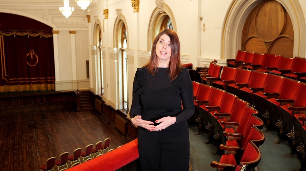 A woman in a black dress standing in an empty theatre speaking to the camera. Red chairs and a curtained stage can be seen in the background.