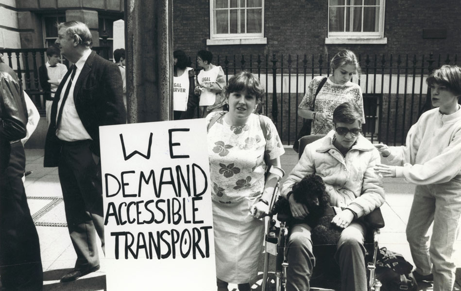 A woman holding a placard that reads ‘We demand accessible transport’ standing alongside a man in a wheelchair.