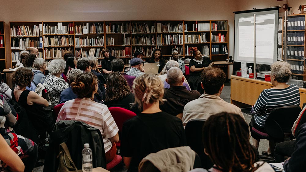 A group of people of a mix of ages and ethnicities attend a talk inside an archive, with shelves of books against the walls