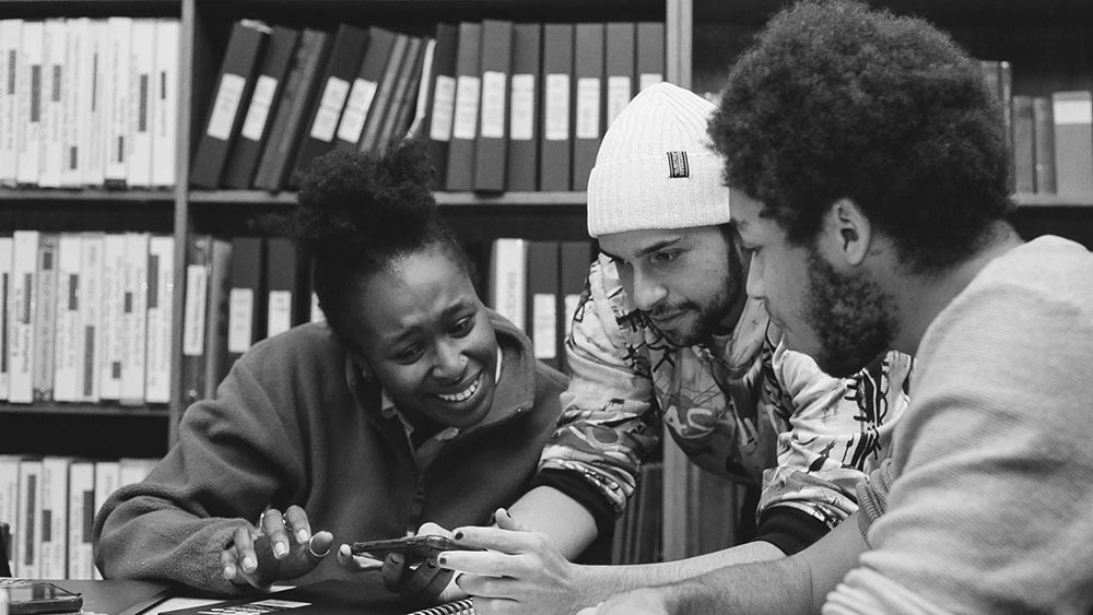 Three people, two of whom have black skin, work together in a room lined with shelves of folders and books