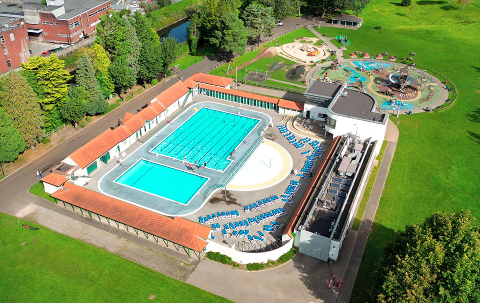 An aerial view of a bright blue swimming pool, set within a park, with a children’s playground located behind it.