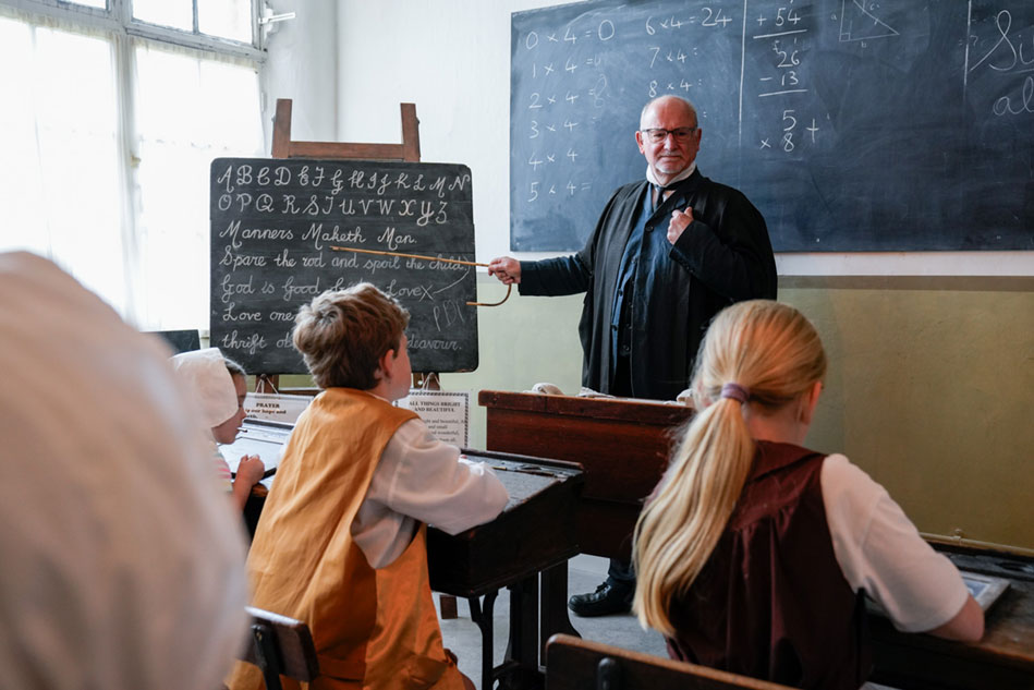 A teacher in period costume standing at a blackboard, demonstrating teaching visiting students at a heritage museum