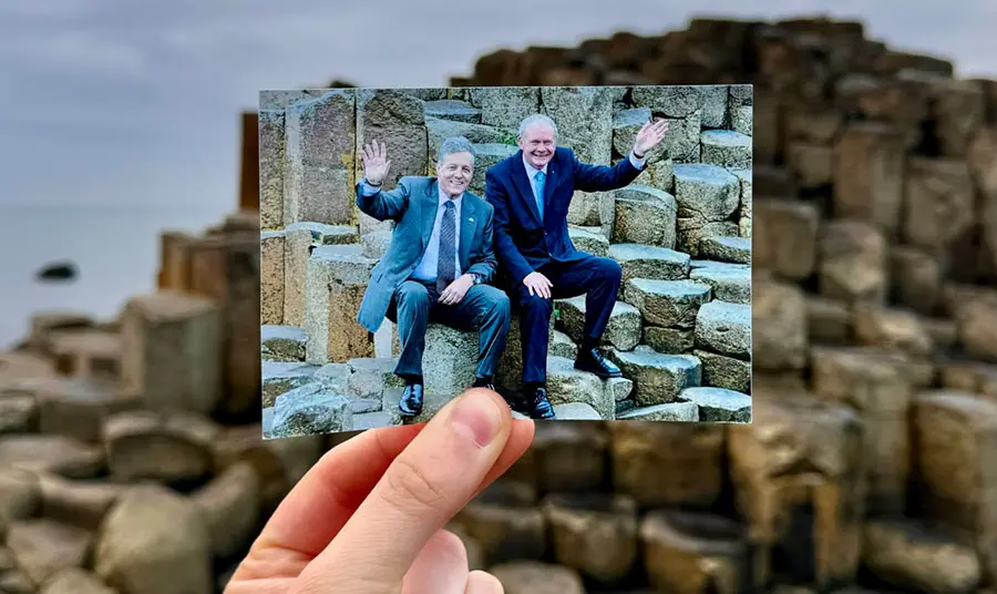 A hand holding a photograph of two men sitting on rock columns, in front of those same rock columns