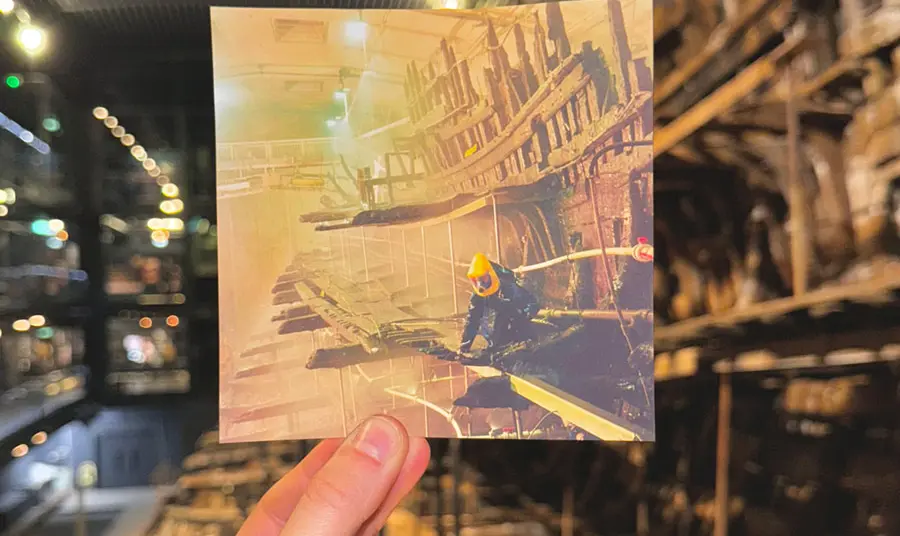 A hand holding a photograph of a person wearing a protective suit among the wreck of a ship, in front of that same wrecked ship