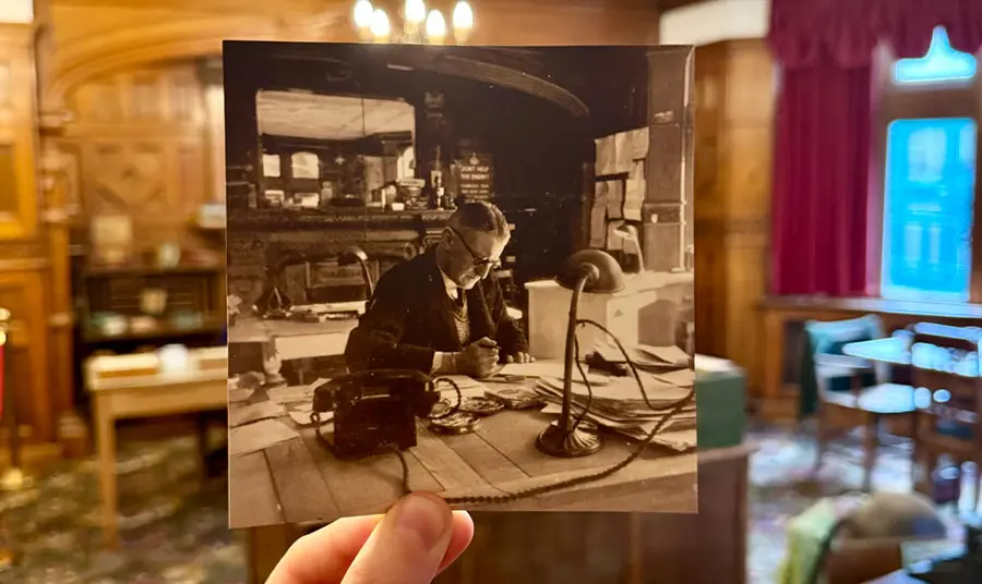 A hand holding a photograph of a man sitting at a desk, in front of that same desk in an office 