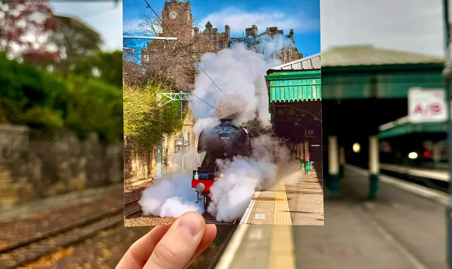 A hand holding a photograph of a train gushing steam as it prepares to pull away from the station, on the platform of that same station