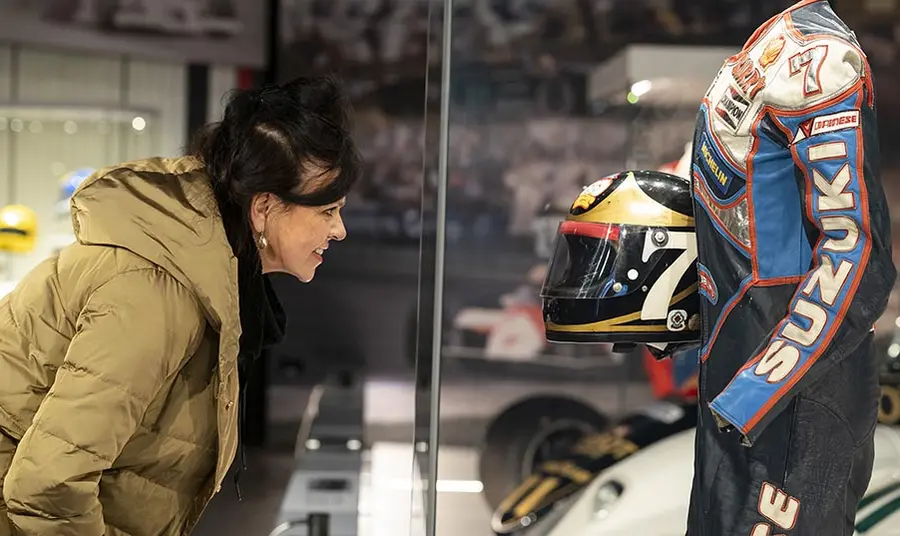 A person with long dark hair wearing a coat looks at a motor-racing helmet and suit