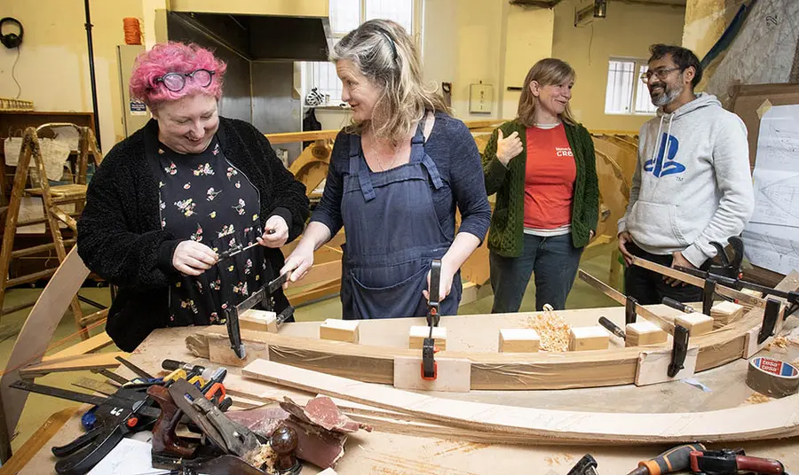 Four people stand talking in front of a table with woodworking equipment