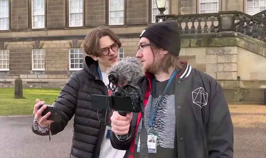 Two young men with cameras and microphone making a film. There is a stately home in the background.