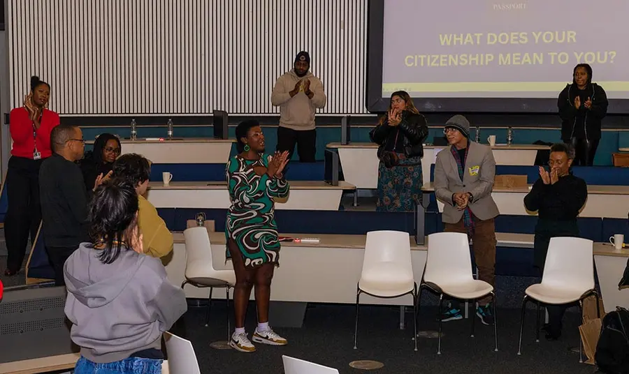 A group of people with black and brown skin involved in a workshop stand in a lecture theatre applauding - the focus of the photo is a black woman wearing a vibrantly patterned dress