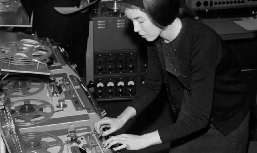 Alt text: A standing woman leant over an audio machine featuring large reels. She is turning a knob with one hand and adjusting a dial with another. 