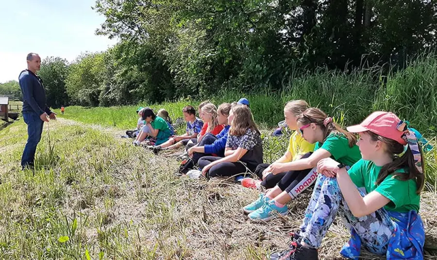 Children sitting on grass at the edge of a woodland listening to an adult