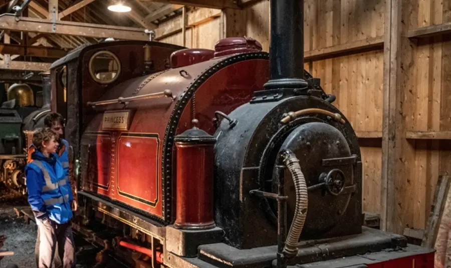 Two visitors investigate a red tank engine in a wooden shed at the Ffestiniog and Welsh Highland Railway.