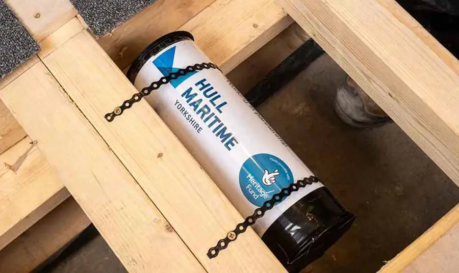 A tube-shaped time capsule with The National Lottery Heritage Fund acknowledgement stamp and Hull Maritime logo secured to wooden flooring beams.