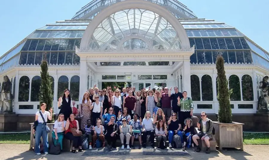 A group of staff outside of glass house on a team away-day, holding up crossed fingers in the style of the National Lottery logo