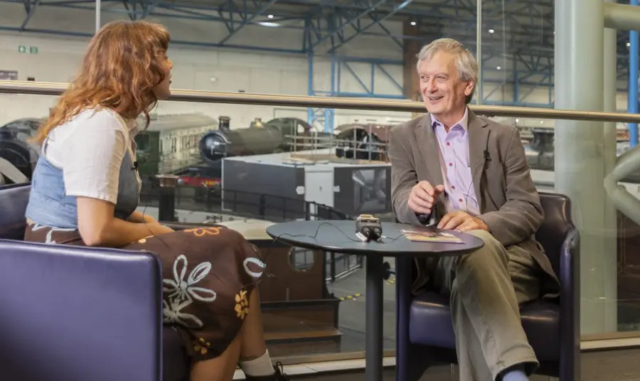 Two people sit at a table in conversation. Behind them are several trains.