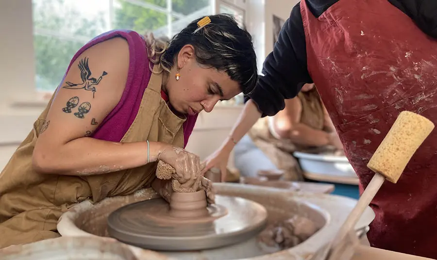 A person in an apron helping another person use a pottery wheel