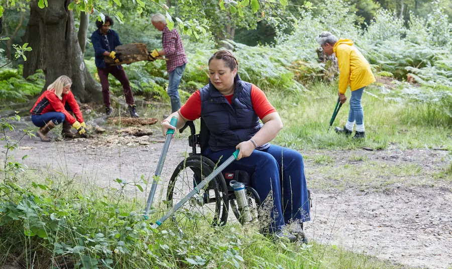 A volunteer in a wheelchair using shears to do practical work in woodland. Other volunteers can be seen doing other practical tasks in the background