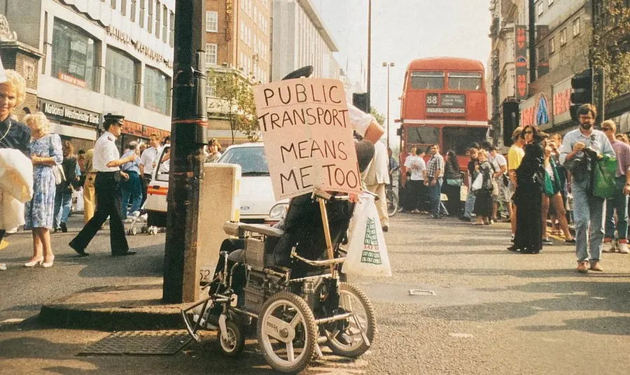 Image of disabled person in 1990s, protesting with plaque saying public transport means me too