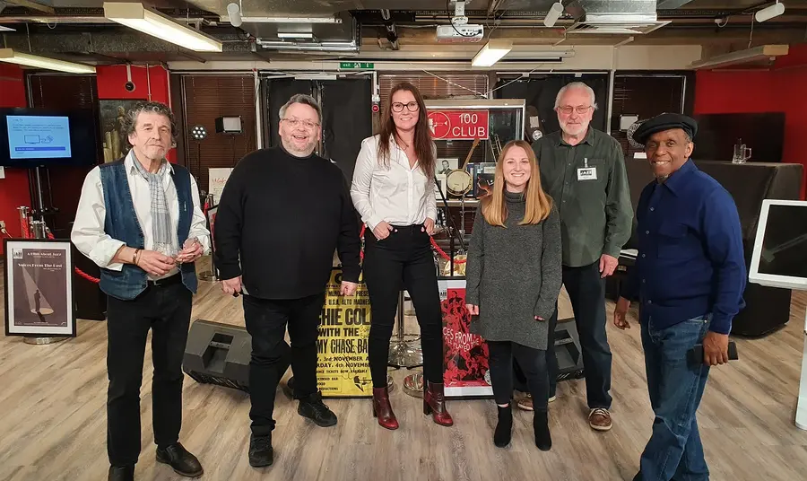 Six people posing for a picture in front of a Jazz music archive, including posters and frames 