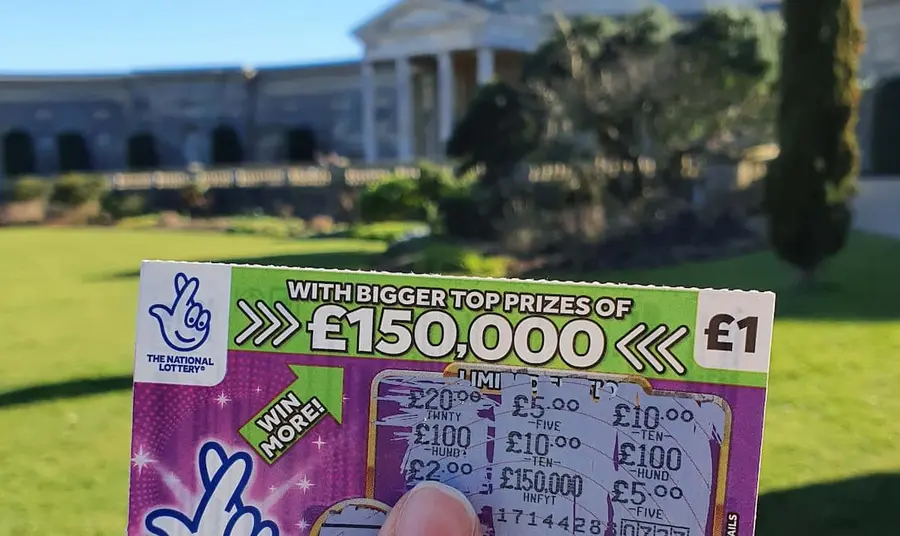 A National Lottery scratchcard held in front of a large building with classical architecture and lawn 