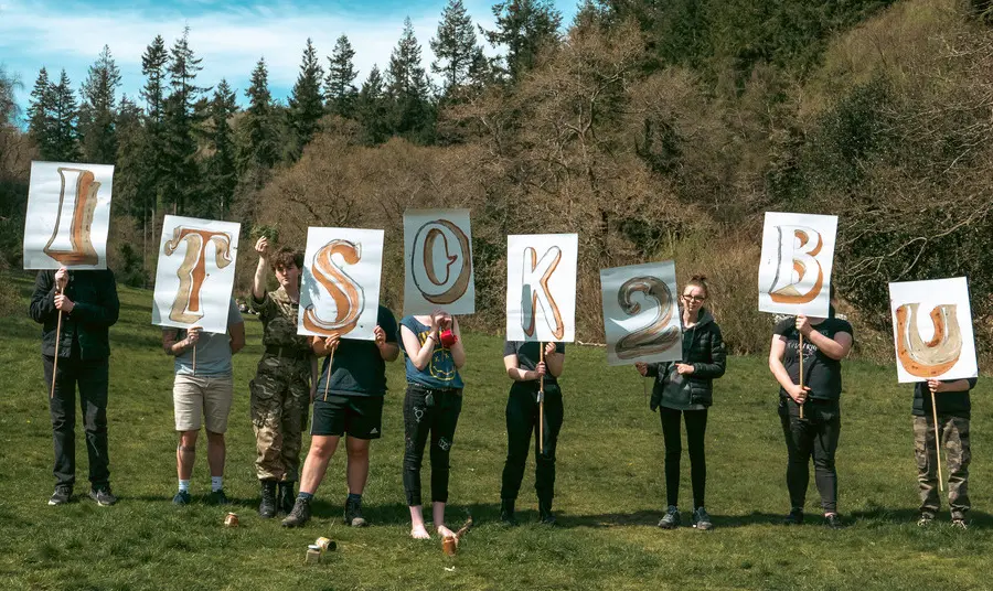 Image of people standing in a line in a countryside setting holding signs reading It's OK 2 B U