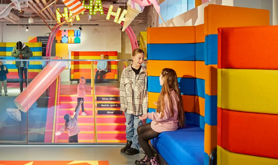 Young children are sat on colourful seating inside the museum, while others are stood on the stairs in the background