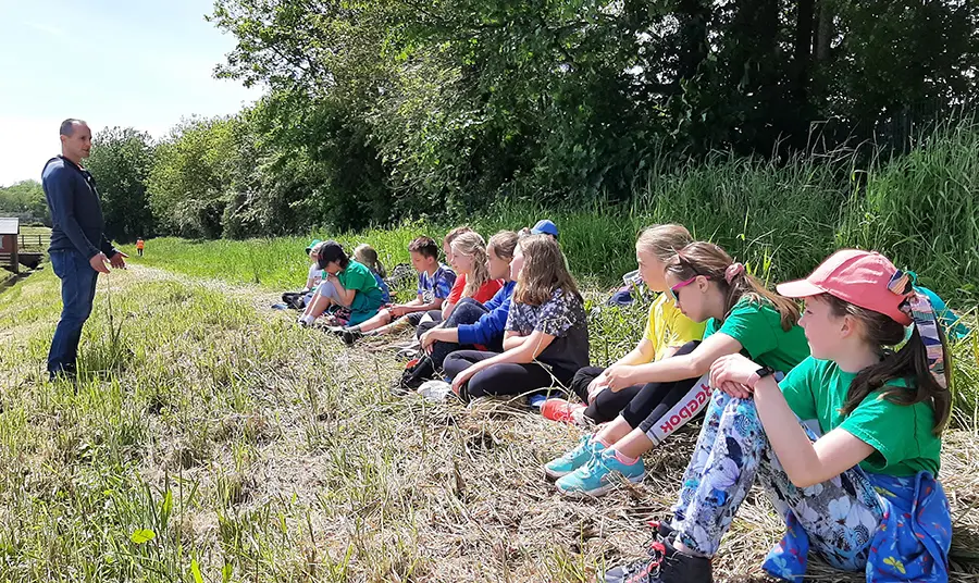 A standing man talking to children sitting on the edge of woodland