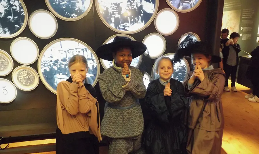 Four children face camera smiling. They are dressed in historic outfits. They are near an exhibition.