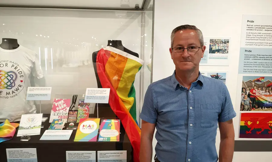 A person standing in front of a glass display of pride flags, t-shirts, leaflets and other memorabilia in a glass case