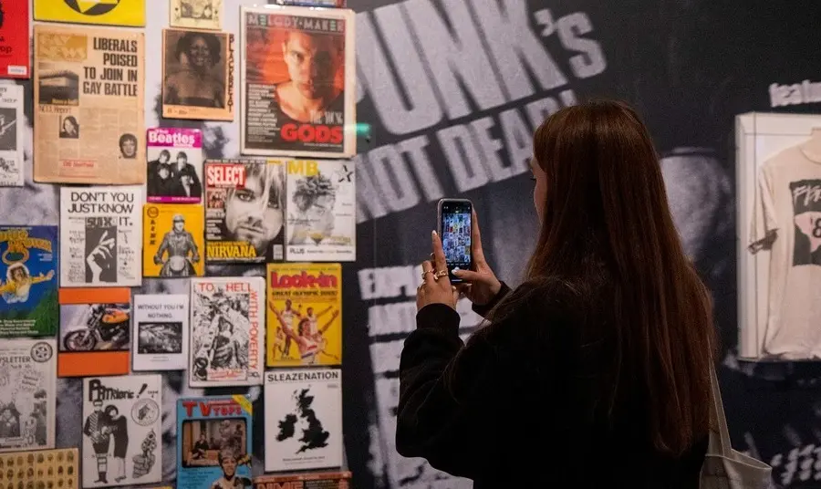 Image of person with long dark hair and dark clothing taking a picture on a mobile of a display of music gig posters at The Museum of Youth Culture