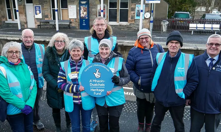A group of people wearing cold weather gear stand on the platform of a rural train station holding a sign