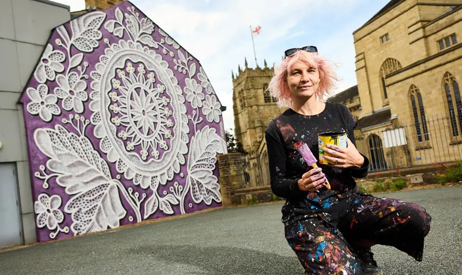 An artist kneels in front of a painted wall with an intricate pattern on a purple background