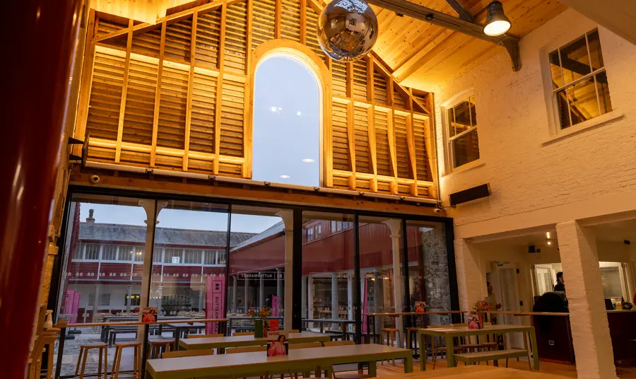 A room with a high vaulted ceiling, a large glitter ball and trestle style tables. The room is warmly lit. Large glass doors look out onto a courtyard.