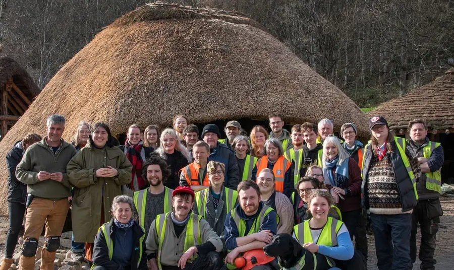 A group of people some wearing high-vis jackets stand together in front of a round house made from wood with a thatch roof