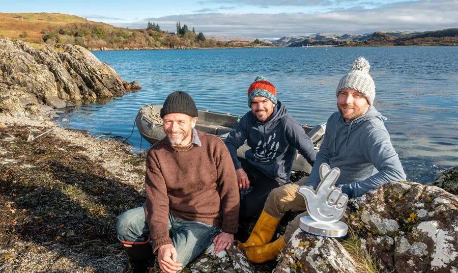 Three people sit on the edge of a loch in Scotland, with a boat in the background and the award in front of them