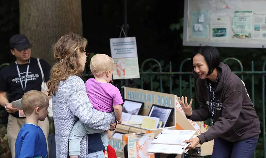 A woman stands outside in front of a bike-mounted display of pictures and other exhibits. She holds a clipboard and talks to a woman and two young children.